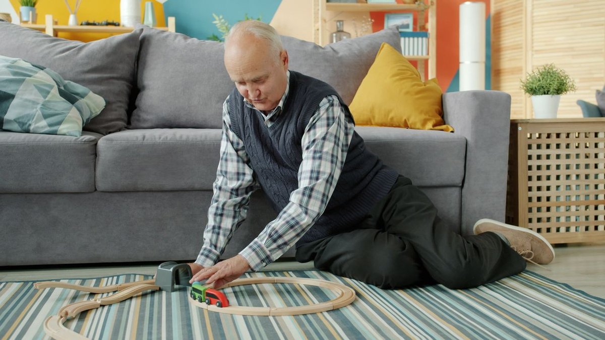Elderly man enjoying time with a wooden train set in a cozy home setting.