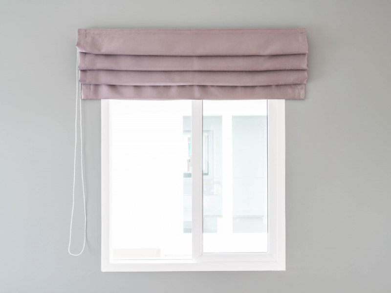 White-framed window with a neatly folded lavender Roman shade mounted above it, featuring horizontal pleats and a white cord hanging on the left side for manual adjustment. The surrounding wall is painted light gray, and through the closed window, a brigh