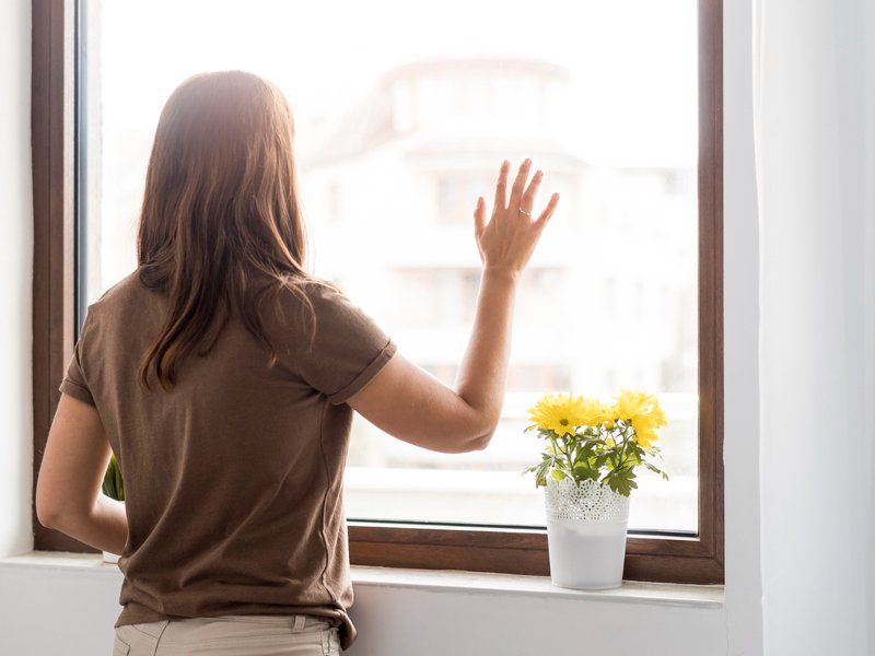Woman with long brown hair wearing a brown shirt and beige pants stands indoors, facing a large window with one hand raised toward the glass. A white pot of yellow flowers sits on the windowsill, and blurred buildings are visible outside. The gesture and