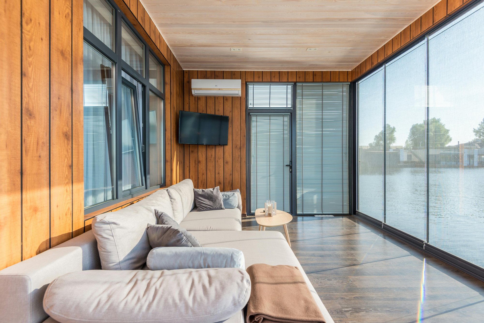 Modern sunroom with floor-to-ceiling glass windows covered by sheer blinds, offering views of water and distant buildings. The room features wood-paneled walls and ceiling, light-colored sofas, a round table with drinks, and a wall-mounted TV and AC unit.