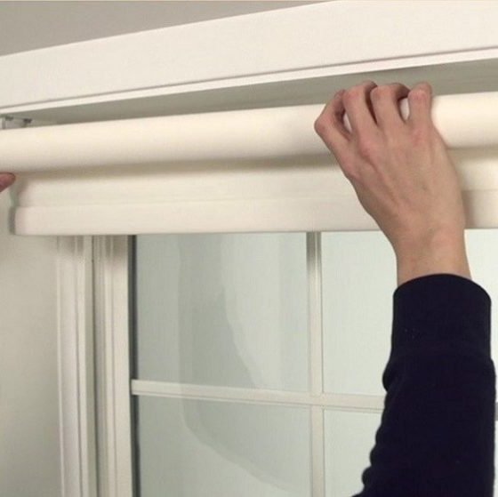 Person installing a white roller shade at the top of a multi-pane window, with hands positioned on either end of the cylindrical roller mechanism. The shade is mounted inside a white window frame, set against a neutral-colored wall.