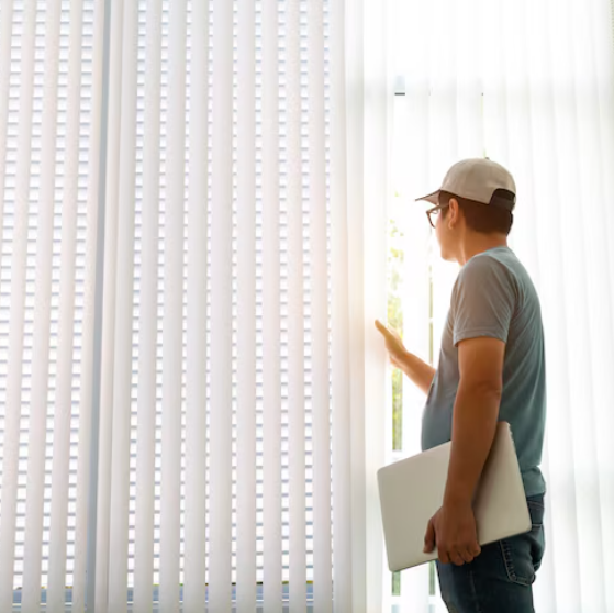 A man standing indoors beside a large window with vertical blinds, gently pulling one aside while holding a closed laptop. Sunlight streams in, creating a warm, contemplative atmosphere.