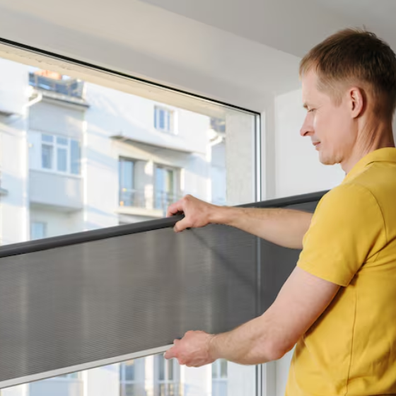Person in a yellow shirt installing a pleated or cellular window blind inside a room. They are adjusting the blind within the window frame, with a multi-story residential building visible outside.