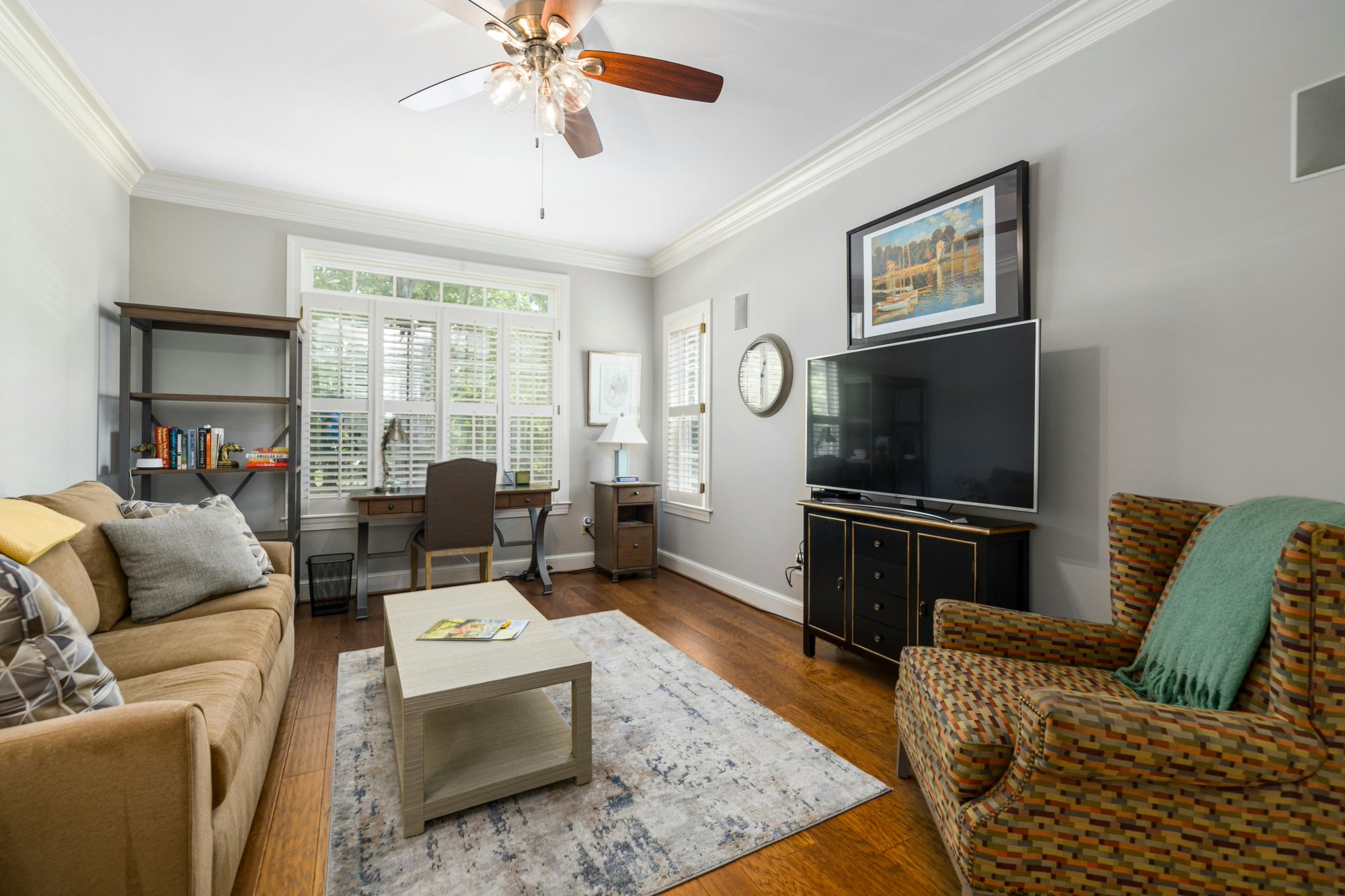 Well-lit living room or home office with hardwood flooring, featuring a brown sofa, patterned armchair with green throw, and a black desk near a window with white plantation shutters.