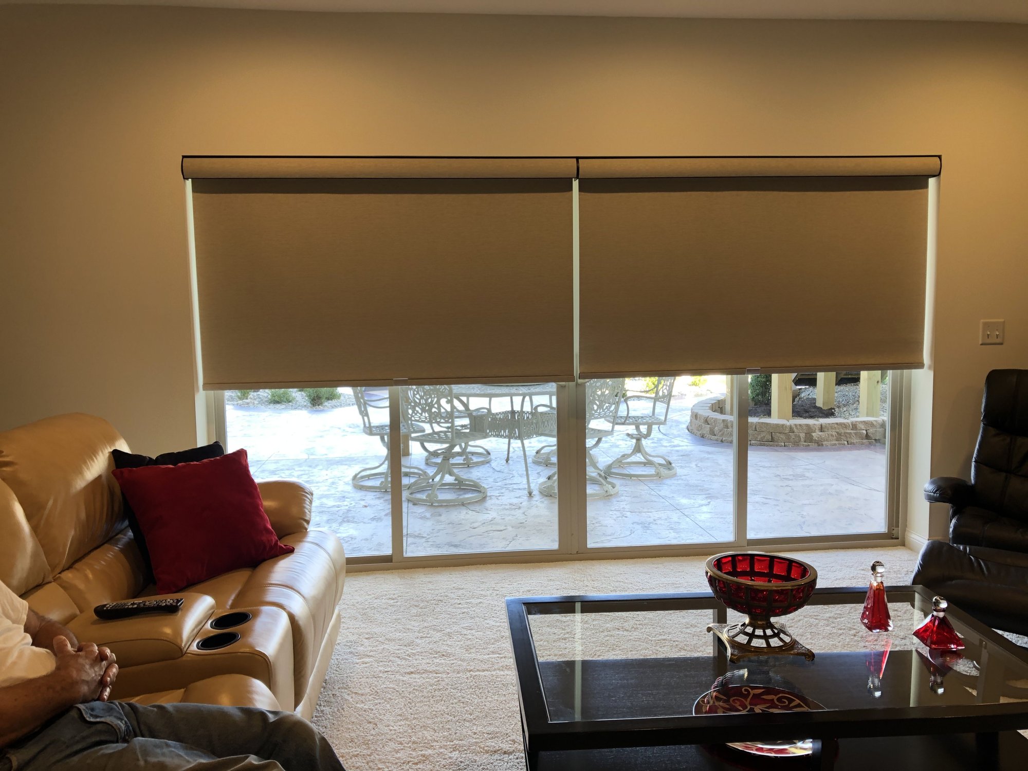 Living room with a large glass sliding door covered by two beige roller shades, allowing natural light to brighten the space. A tan leather sofa with red and black accent pillows sits beside a black leather recliner and a glass coffee table adorned with r