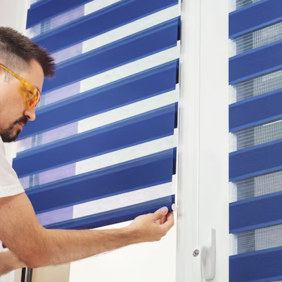 A man installing blue zebra blinds on a white-framed window, adjusting the mechanism while wearing yellow-tinted safety glasses.