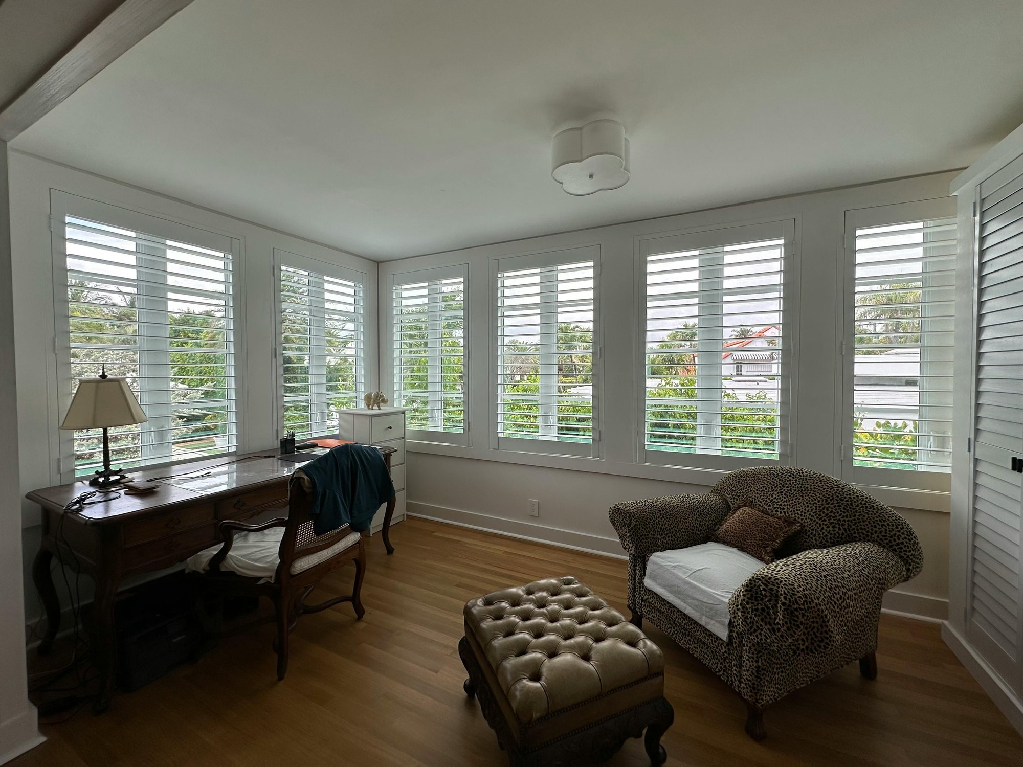 Cozy, sunlit room with large windows fitted with white plantation shutters, allowing filtered light and views of greenery outside. The space features wooden flooring, white walls, a vintage desk with a lamp and chair, and a leopard-print armchair with a w
