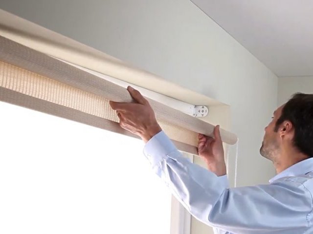 Person adjusting a light-colored, semi-transparent roller shade by holding its bottom edge with both hands near a window frame. The roller mechanism is mounted at the top of the window, and natural light filters through the shade.