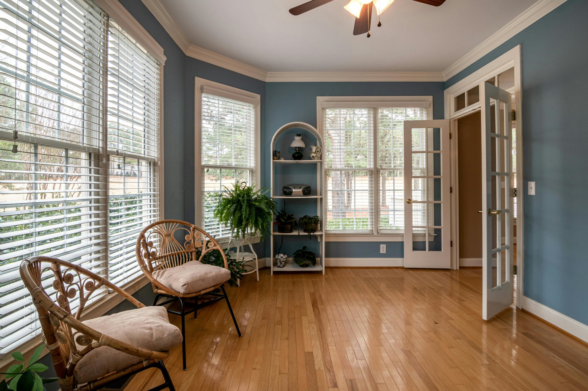 Bright sunroom with soft blue walls, polished hardwood floors, and large windows that fill the space with natural light. Two rattan chairs with beige cushions sit near the windows, creating a cozy seating area. A white arched shelving unit holds potted pl