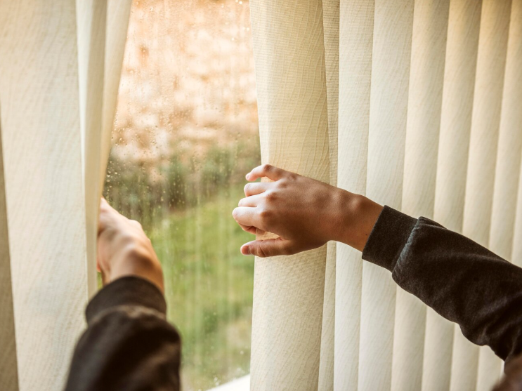 Close-up of two hands parting light-colored vertical blinds to look outside through a window. The person, wearing a dark long-sleeve shirt, reveals a blurred view of greenery and a beige surface.