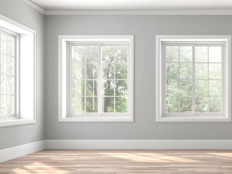 Bright, empty room with light gray walls, white trim, and light wood flooring. Three large multi-pane windows with white frames let in natural light and offer views of lush greenery outside.