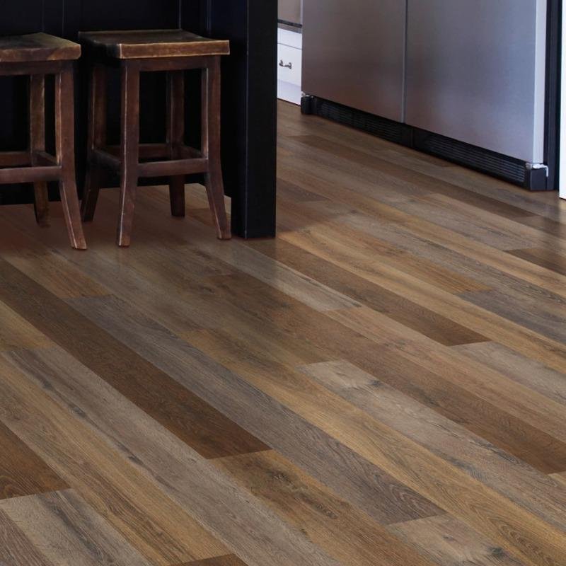 Close-up of a modern kitchen or dining area floor featuring staggered wood-look planks in varying shades of brown. The textured grain adds warmth and visual depth. Two wooden bar stools with square seats are visible in the upper left corner, while part of