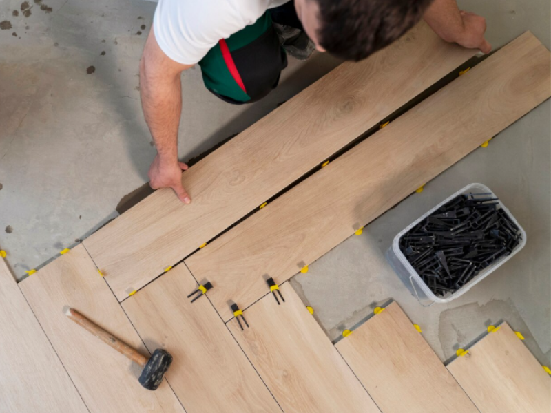 Person installing laminate flooring with yellow spacers, using tools to align planks over a concrete subfloor.