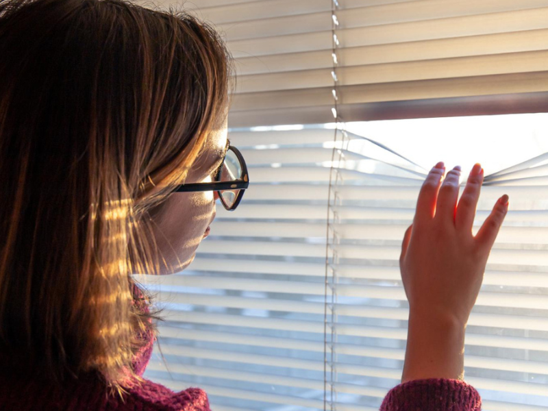 A woman in glasses and maroon sweater peeking through horizontal window blinds, gently lifting one slat to look outside. Sunlight filters through the blinds, casting soft shadows and highlights across the person's face and clothing
