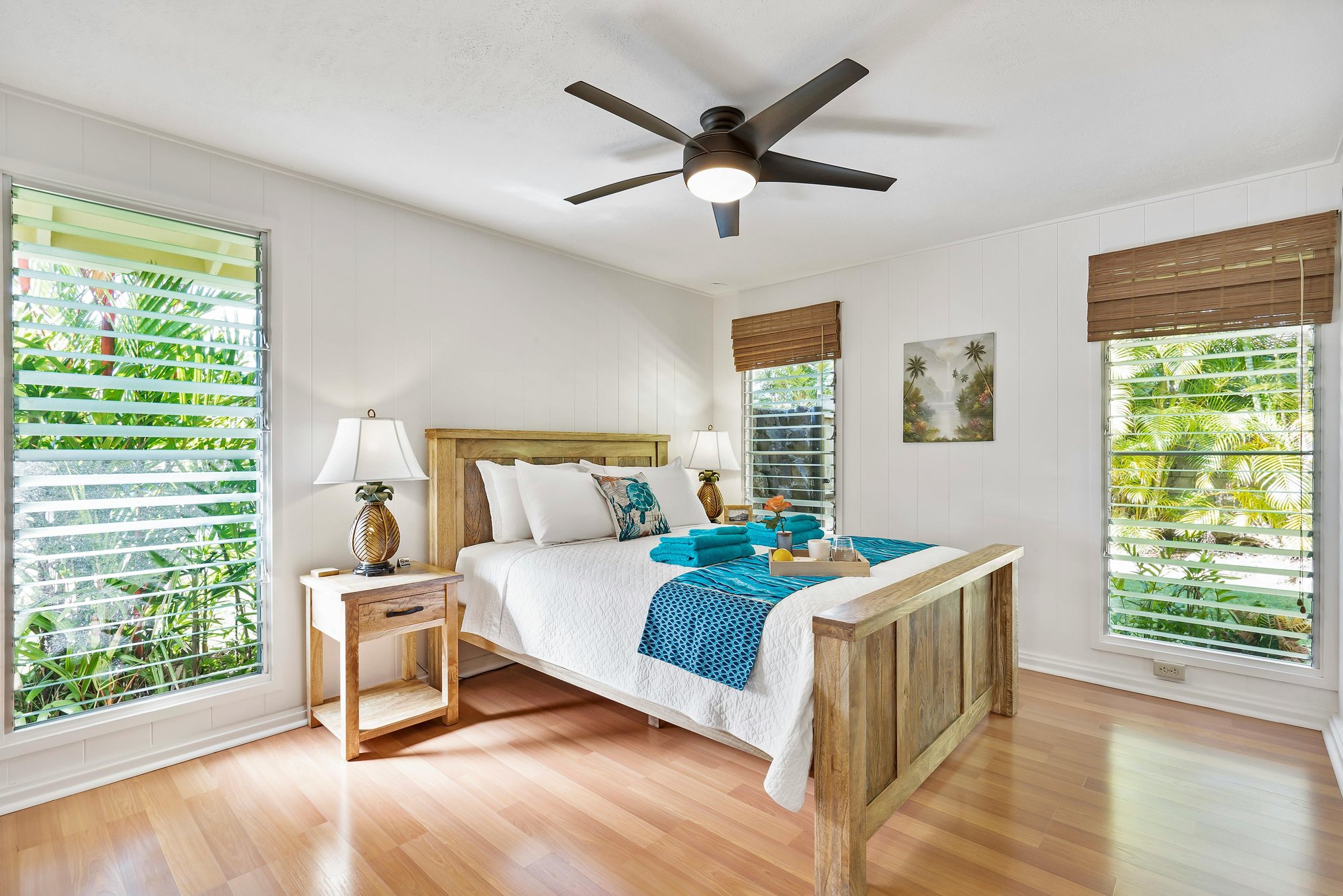 Bright tropical-themed bedroom with white bedding, turquoise accents, pineapple lamps, and light wood laminate flooring. Three large windows with horizontal blinds let in natural light and offer views of lush greenery outside.