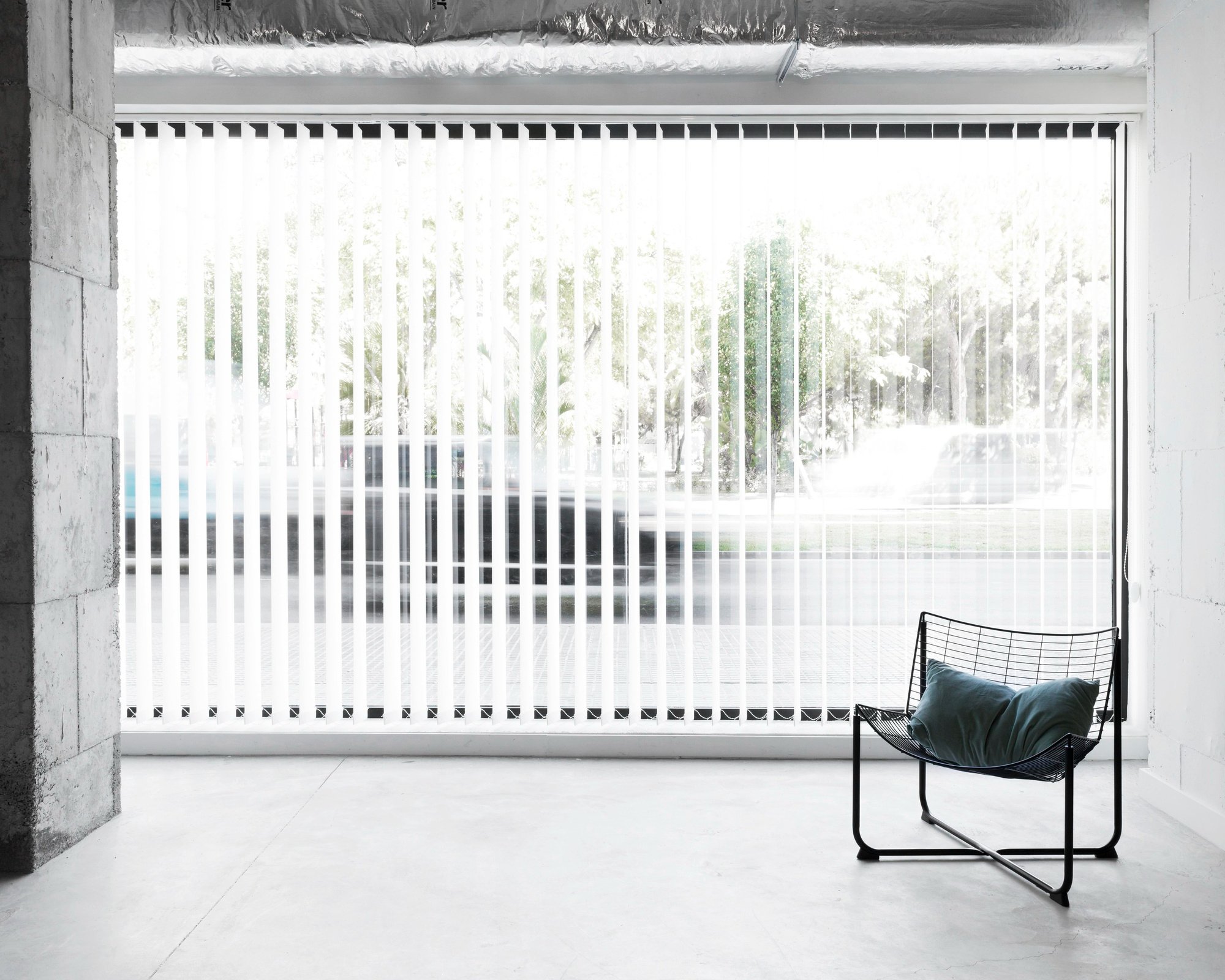 Minimalist interior with a floor-to-ceiling window covered by vertical blinds. A black metal-framed chair with a teal cushion sits on a light gray concrete floor. Sunlight filters through the blinds