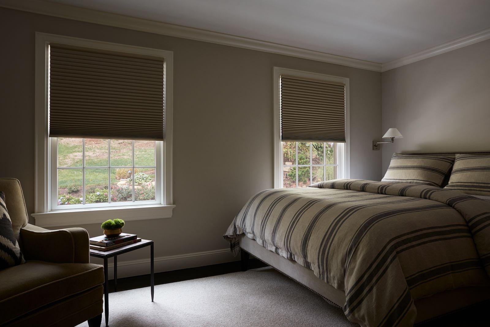 Modern bedroom with striped neutral bedding, light gray walls, and two windows covered by closed beige blinds. Soft natural light filters through, revealing a grassy outdoor view. A beige armchair and side table with books and a potted plant sit beside th