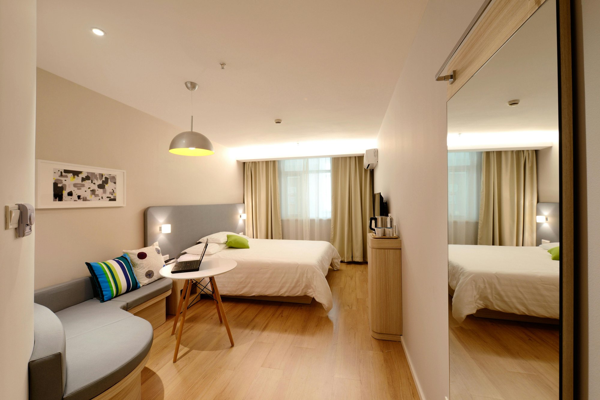 Modern hotel room with two beds featuring white bedding, a small round table with a laptop and cup, and a cushioned bench with colorful pillows beneath abstract artwork. A pendant light hangs above the table, and a large mirror reflects the room. Sheer an