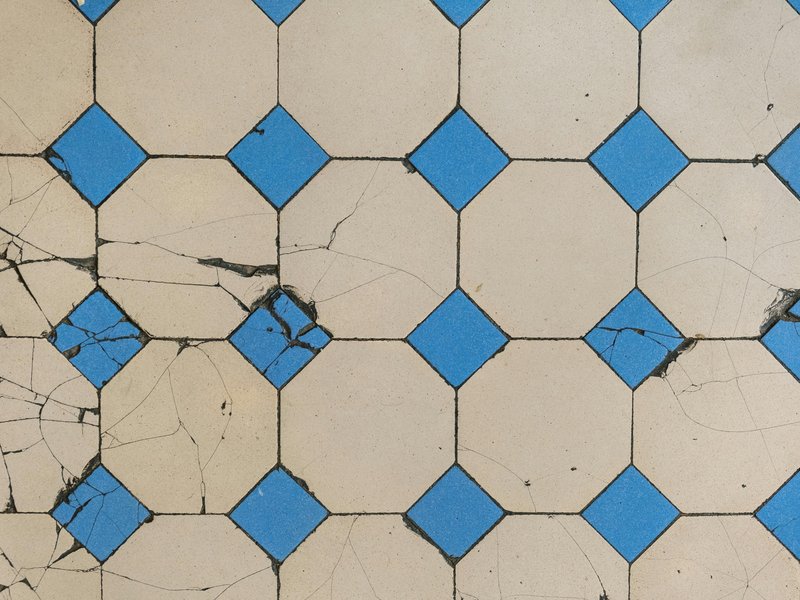 Tiled floor with a geometric pattern of large beige square tiles and smaller blue tiles forming diamond shapes at the corners. Several tiles in the lower left area are cracked, with irregular lines disrupting the symmetry.