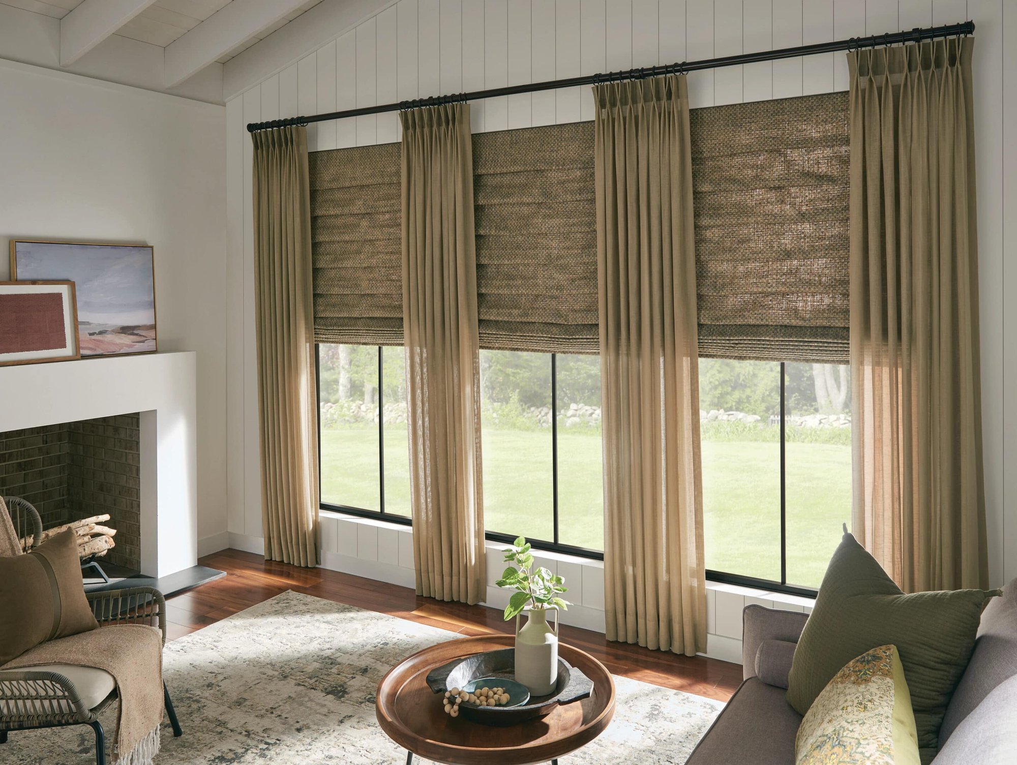 Cozy living room with large windows dressed in woven Roman shades and sheer beige curtains, offering a view of a green lawn and garden. A gray sofa with patterned cushions sits in front of the windows, paired with a round wooden coffee table holding a pot