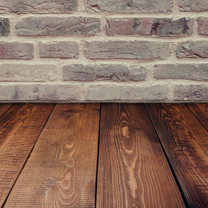 Rustic interior with rich brown wooden plank flooring and a textured brick wall in shades of red, pink, and gray. The contrast between the smooth wood grain and rough brick surface creates a visually striking, warm atmosphere—ideal for showcasing interior
