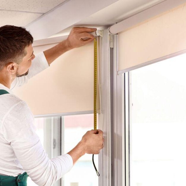 Person in green overalls and a white shirt measuring the height of a window frame with a yellow tape measure. The window has a partially rolled-up roller blind, and the background shows a light-colored wall and white tiled ceiling.