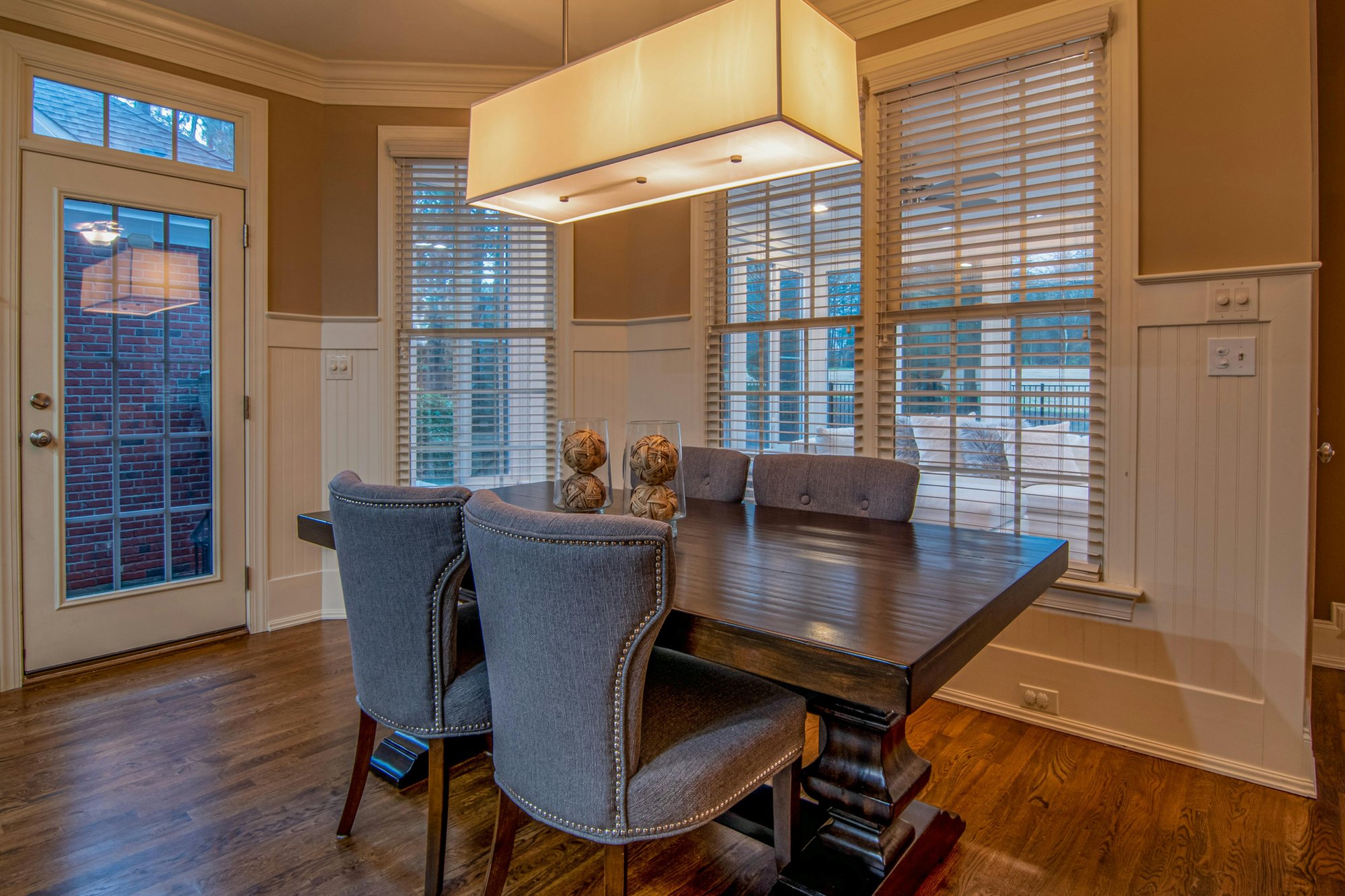 Elegant dining area with a dark wooden table surrounded by six tufted gray-blue chairs with nailhead trim. A cream-colored rectangular pendant light hangs above the table, which is decorated with three natural-textured spherical sculptures. The room featu