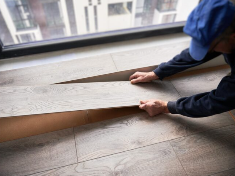 Person installing light wood-look laminate flooring near window, aligning planks for precise fit in a bright interior space.