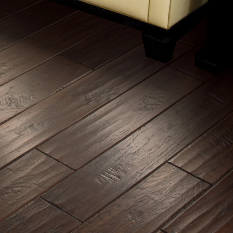Close-up of a dark wooden floor with rich grain patterns and staggered planks featuring beveled edges for added depth. In the upper right corner, part of a cream-upholstered furniture piece with black legs is visible, creating a striking contrast against