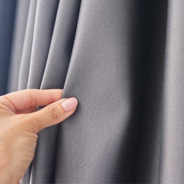 Close-up of a hand gently inspecting a thick, dark gray curtain, revealing its smooth texture and soft folds. The fabric appears suitable for blackout or thermal insulation, making it ideal for enhancing privacy and energy efficiency in interior spaces.