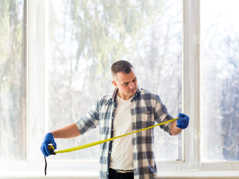 Person wearing a plaid shirt, white t-shirt, and blue gloves stands indoors in front of large windows, holding a yellow measuring tape. They appear to be measuring the window width, with a blurred view of trees visible outside.
