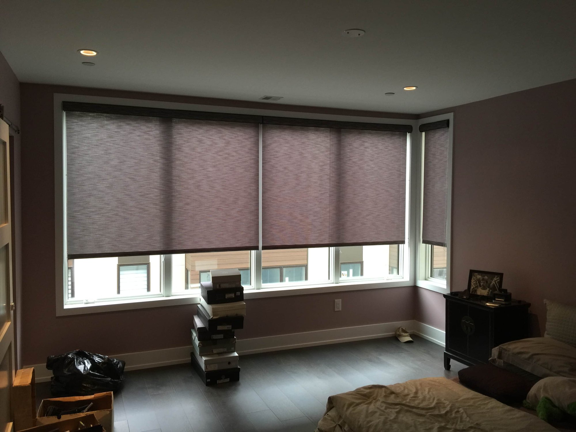 Modern bedroom with large windows covered by four partially lowered roller shades, allowing soft natural light to enter. The walls are painted muted purple, and the dark wood flooring adds warmth. A bed with rumpled bedding sits in the lower right corner,