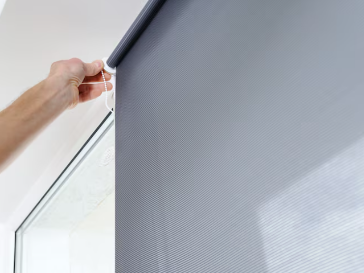 Close-up of a hand adjusting a dark gray roller blind using a beaded chain mechanism. The textured, semi-opaque fabric is mounted inside a window frame, demonstrating manual operation. The lack of full opacity suggests limited light blocking, making it le