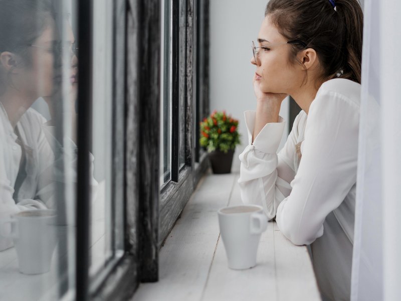 Person in glasses and a white long-sleeved shirt sits indoors, resting their chin on their hand while gazing thoughtfully out a window. A white coffee mug and a small potted plant with red and green foliage sit on the windowsill. The window reflects the p