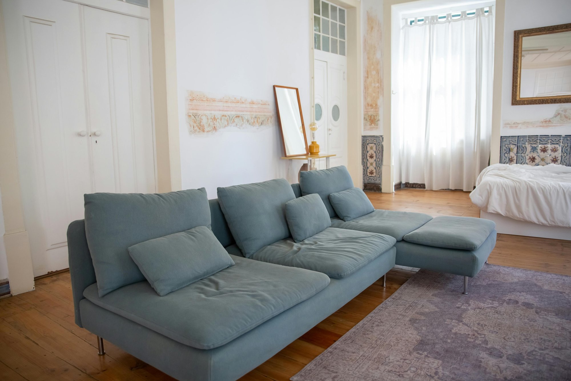 Stylish living space with a muted teal sectional sofa on a light purple rug over wood flooring. A bed with white bedding sits near sheer curtains that let in soft natural light.
