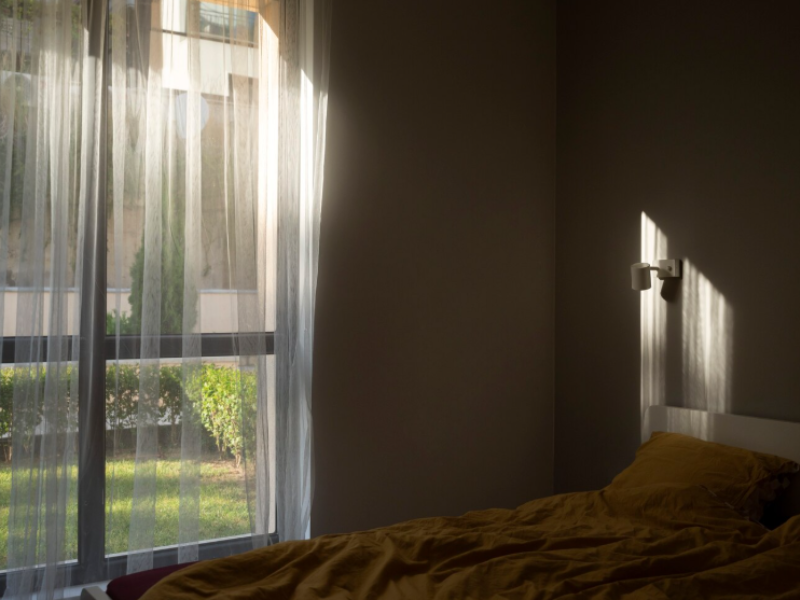 Sunlit bedroom with sheer white curtains covering a large window, casting soft shadows on the wall and illuminating a bed with mustard-colored bedding. Outside, a manicured lawn and building facade are visible. A small wall-mounted light fixture sits abov