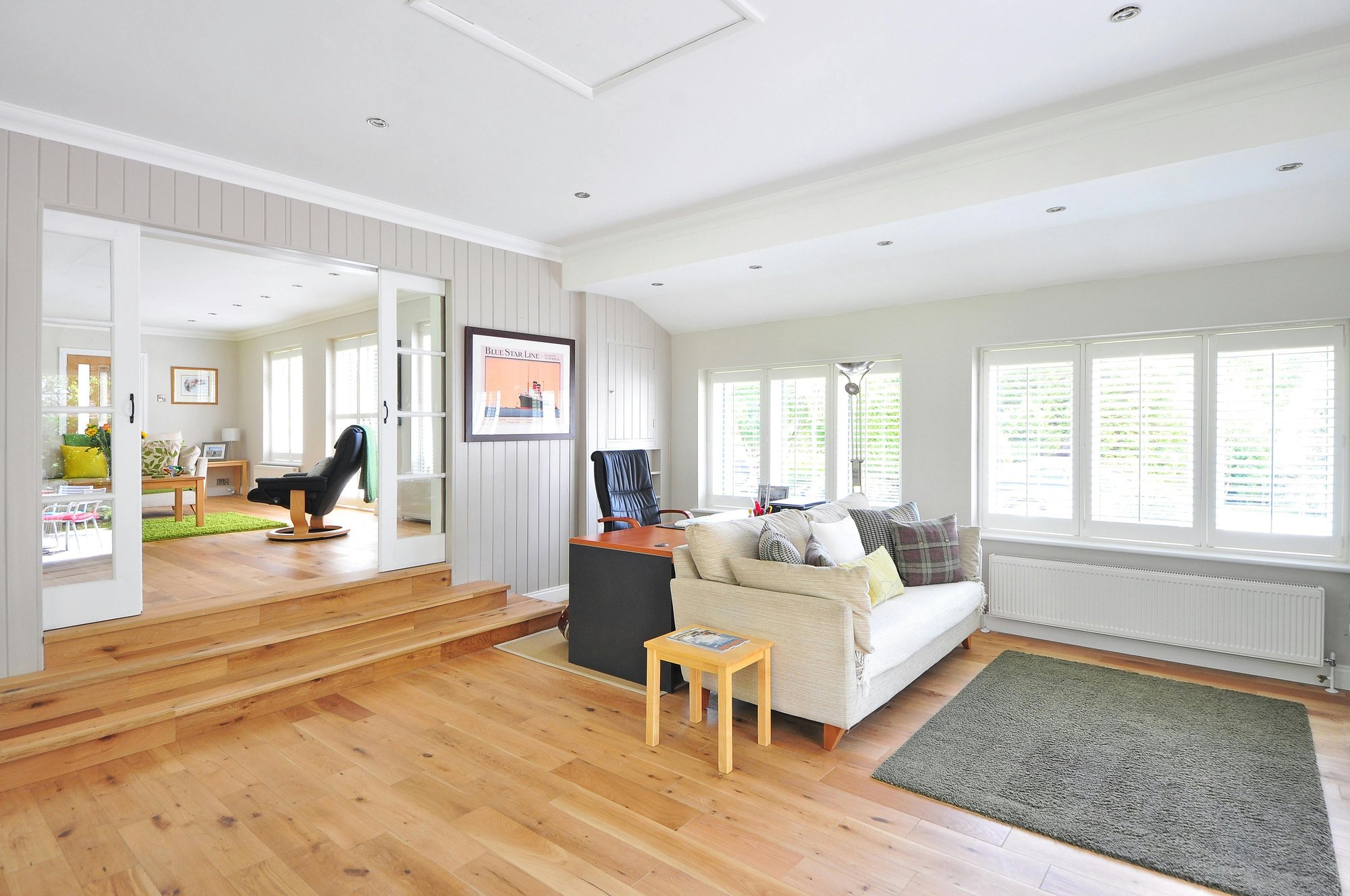 Bright, open-plan living area with light-colored walls and wooden flooring. A beige sofa with cushions sits on a green rug beside a small wooden side table. Behind the sofa, a black office chair and desk are positioned near large white-framed windows with