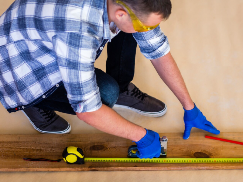 Person in plaid shirt and gloves measures a wooden plank with tape and square tool, prepping for precise DIY work.