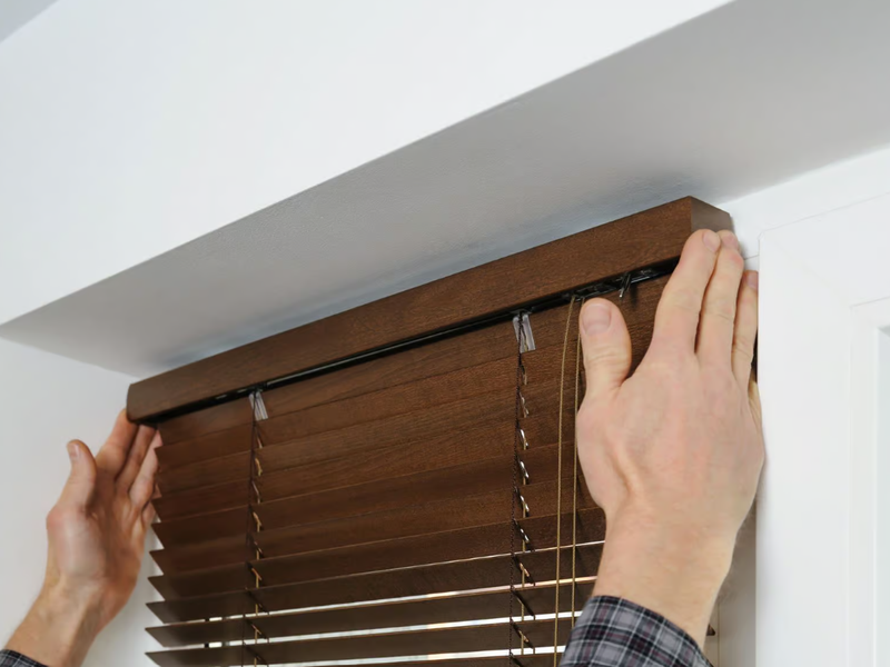 Close-up of a person in a plaid shirt installing brown horizontal blinds inside a white window frame. Their hands are positioned on either side of the top rail, securing or aligning the fixture. The blinds appear to be made of wood or faux wood, adding a